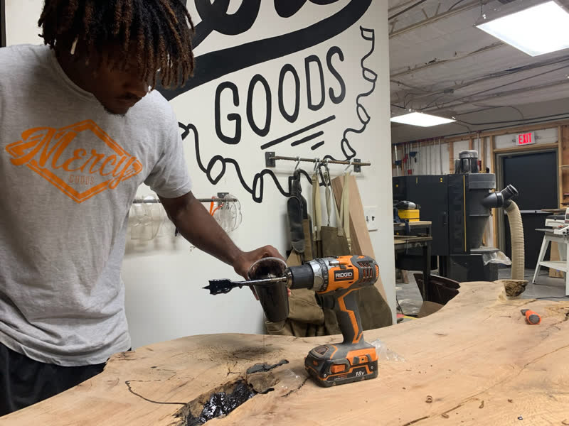 A young maker in a Mercy Goods shirt mixing epoxy with a drill paddle over a live-edge slab