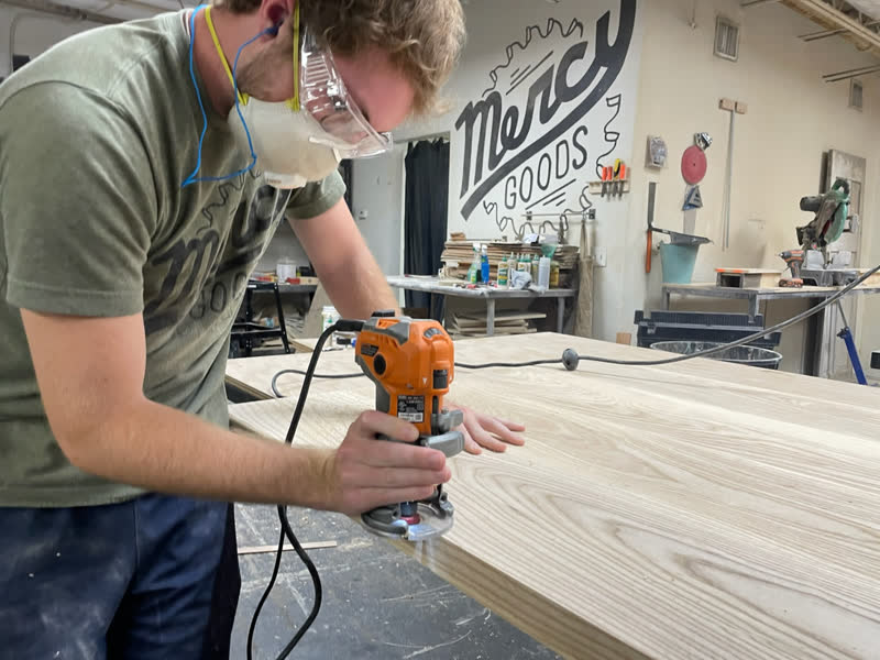 A maker in a respirator and safety glasses easing an edge with a trim router