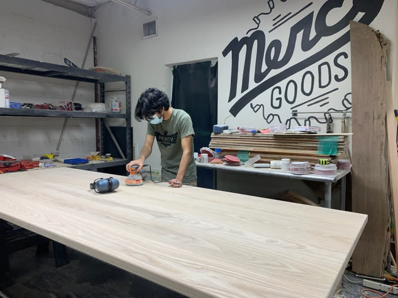 A maker in a Mercy Goods shirt sanding a wide tabletop in front of the painted mural