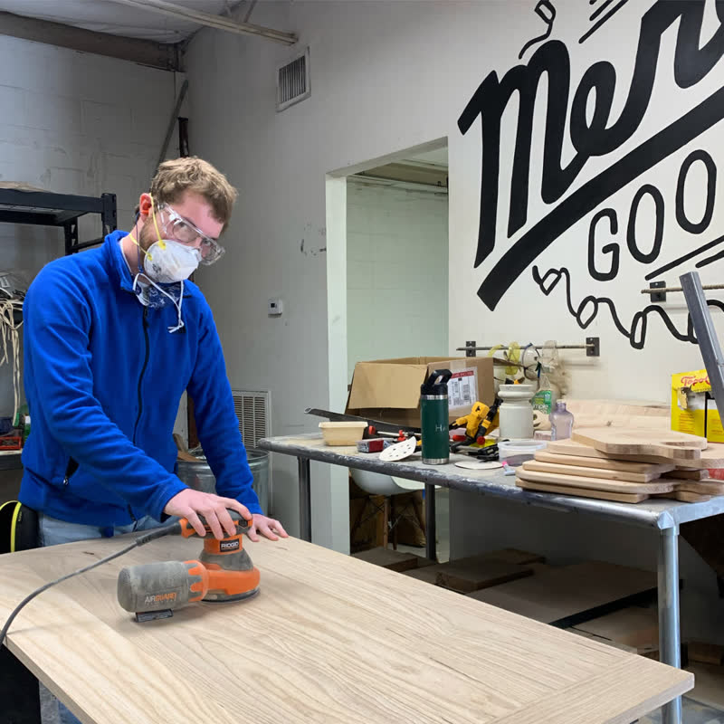 A young maker in safety glasses sanding a countertop beside the Mercy Goods mural