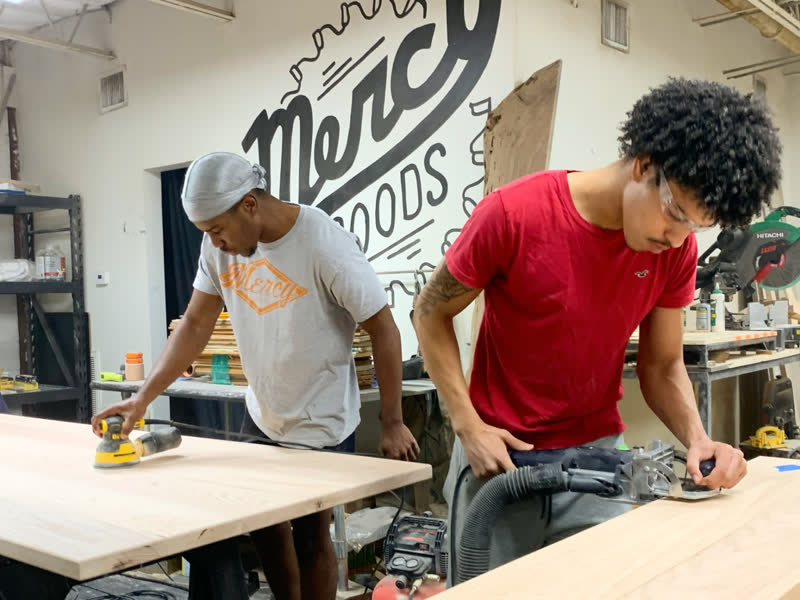 Two young makers sanding and routing tabletops beneath the Mercy Goods mural