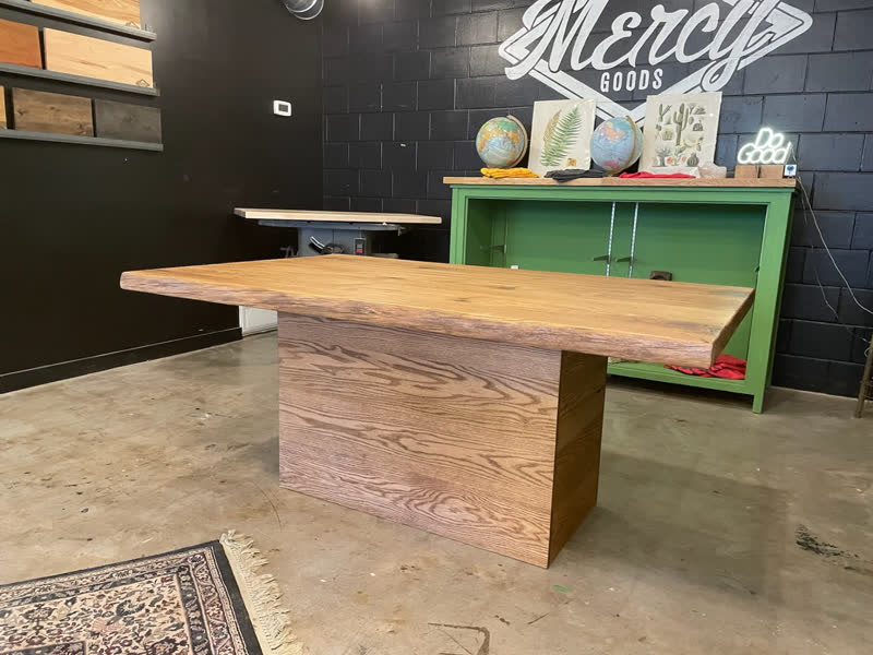 A live-edge table on an oak pedestal base beneath the painted Mercy Goods mural