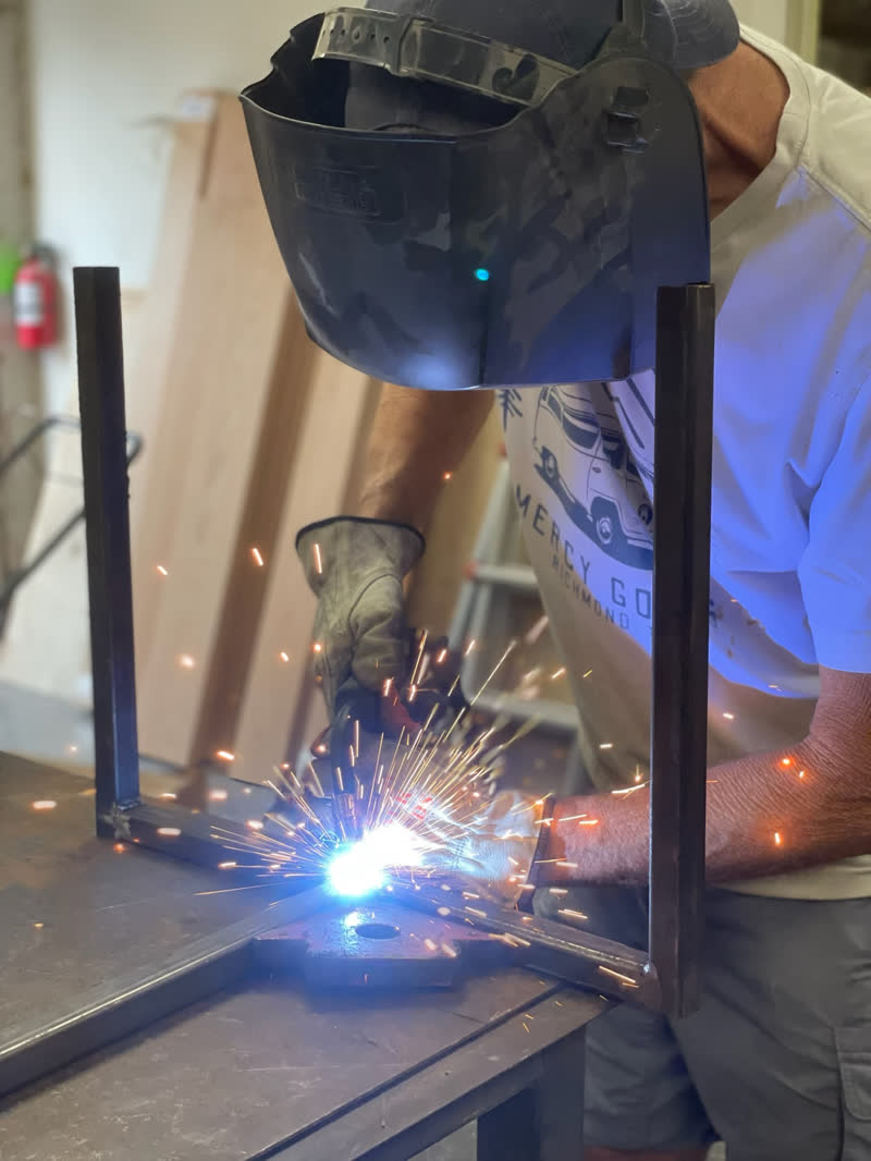 A maker in a Mercy Goods shirt welding sparks into a steel table frame