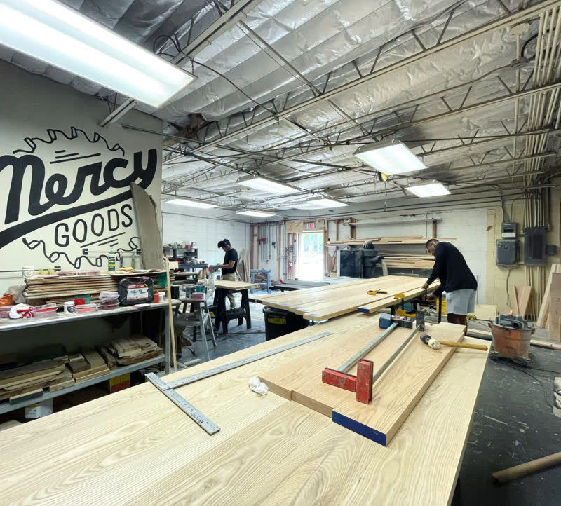 The full Mercy Goods workshop with the painted Mercy logo on the wall and makers at the benches