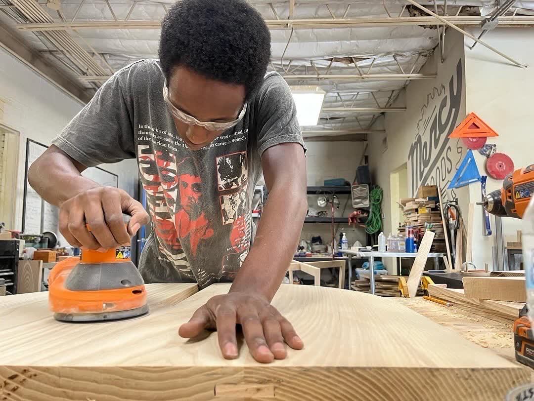 A young maker sanding a wide board in the Mercy Goods shop, with the Mercy Goods mural behind him